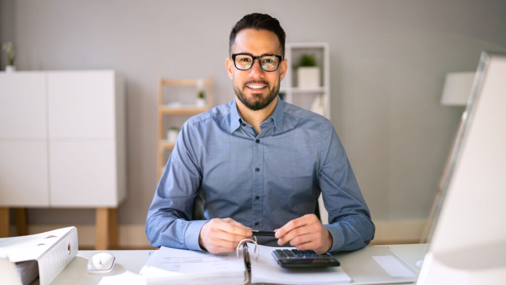 Contador branco, de camisa social e óculo preto sorrindo em um escritório, segurando uma caneta e sentado em uma mesa.