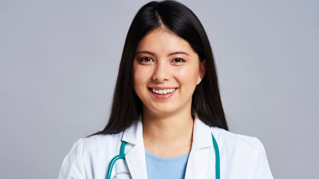 Médica nova sorrindo e olhando para frente, com jaleco e estetoscópio azul no pescoço.